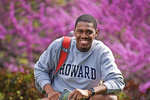 Student seated outside near flowers with a book bag, holding a text book and smiling
